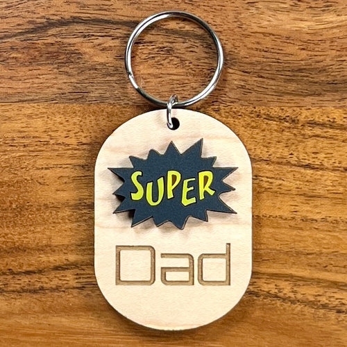 Super Dad laser-cut maple wood keychain with black and yellow acrylic comic burst and engraved text.