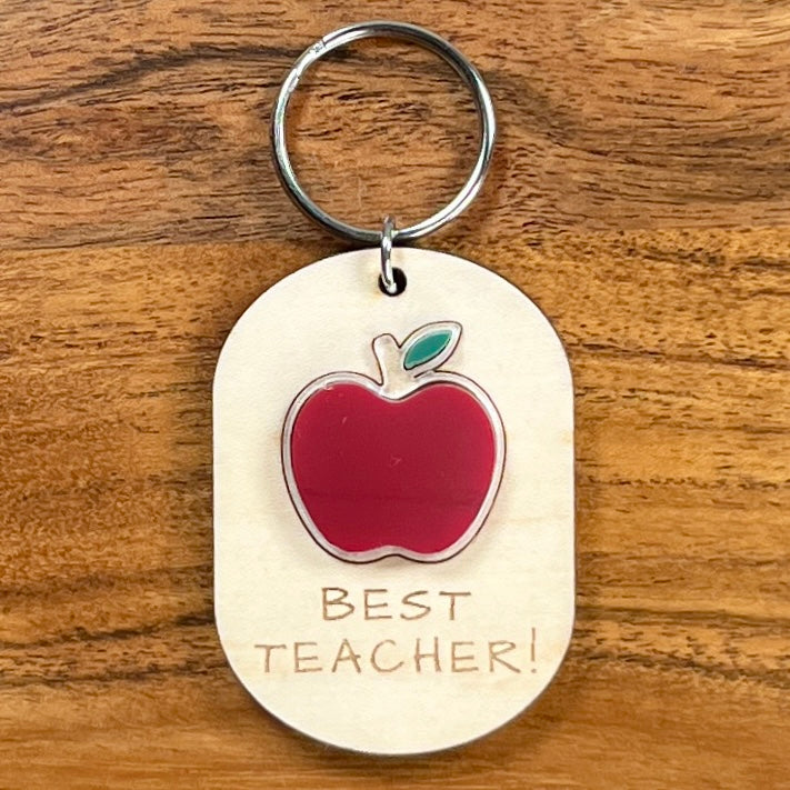 Oval wooden keychain with red and green acrylic apple, engraved 'Best Teacher!' on a wood surface with a metal split ring. 