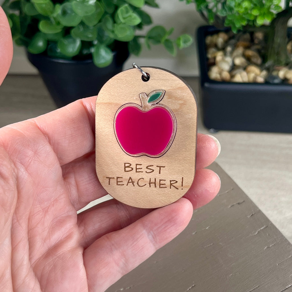 A hand holding an oval wooden keychain with a layered red and green acrylic apple and the engraving 'Best Teacher!' 