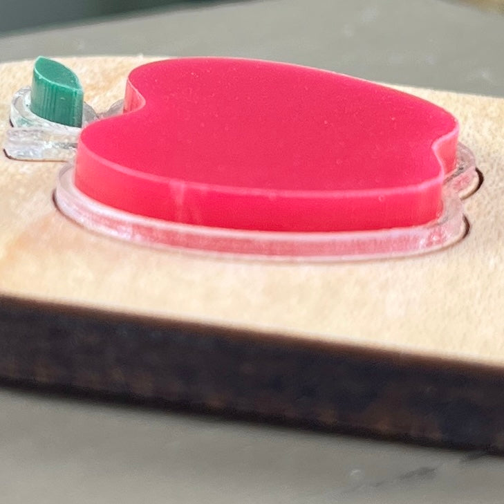 Side view of an oval wooden keychain with a layered red and green acrylic apple, highlighting the 3D effect of the design.