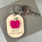 Oval wooden keychain with red and green acrylic apple, engraved 'Best Teacher!' attached to a metal key on a brown surface. 