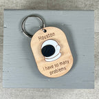 Oval wooden keychain with white and black acrylic space helmet, engraved 'Houston, I have so many problems,' on a gray surface. 