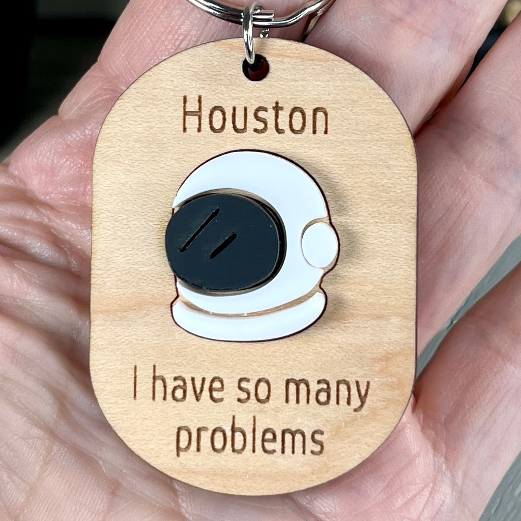 A hand holding a wooden keychain with a white and black acrylic space helmet, engraved 'Houston, I have so many problems.'