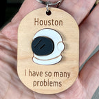 A hand holding a wooden keychain with a white and black acrylic space helmet, engraved 'Houston, I have so many problems.'