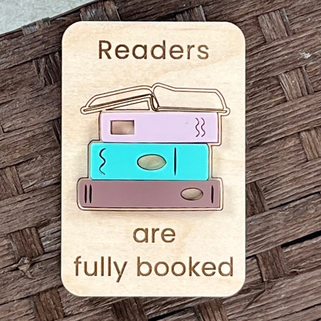 A rectangular wooden magnet with rounded corners featuring the engraved text 'Readers are fully booked' and an acrylic design of three stacked books in pastel colors, placed on a dark woven wood surface.