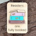 A rectangular wooden magnet with rounded corners featuring the engraved text 'Readers are fully booked' and an acrylic design of three stacked books in pastel colors, placed on a dark woven wood surface.
