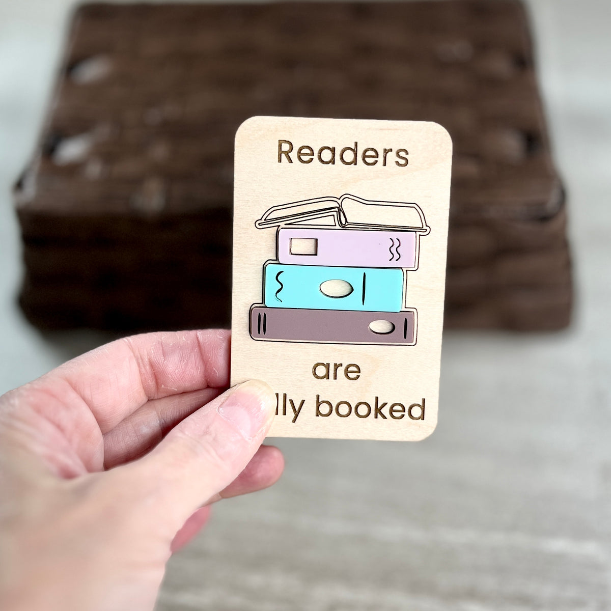 A hand holds a small wooden magnet that reads 'Readers are fully booked' with a simple acrylic stacked book design in pastel colors. The background shows a blurred stack of similar wooden cards on a light surface.
