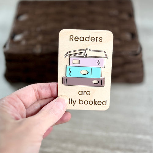 A hand holds a small wooden magnet that reads 'Readers are fully booked' with a simple acrylic stacked book design in pastel colors. The background shows a blurred stack of similar wooden cards on a light surface.