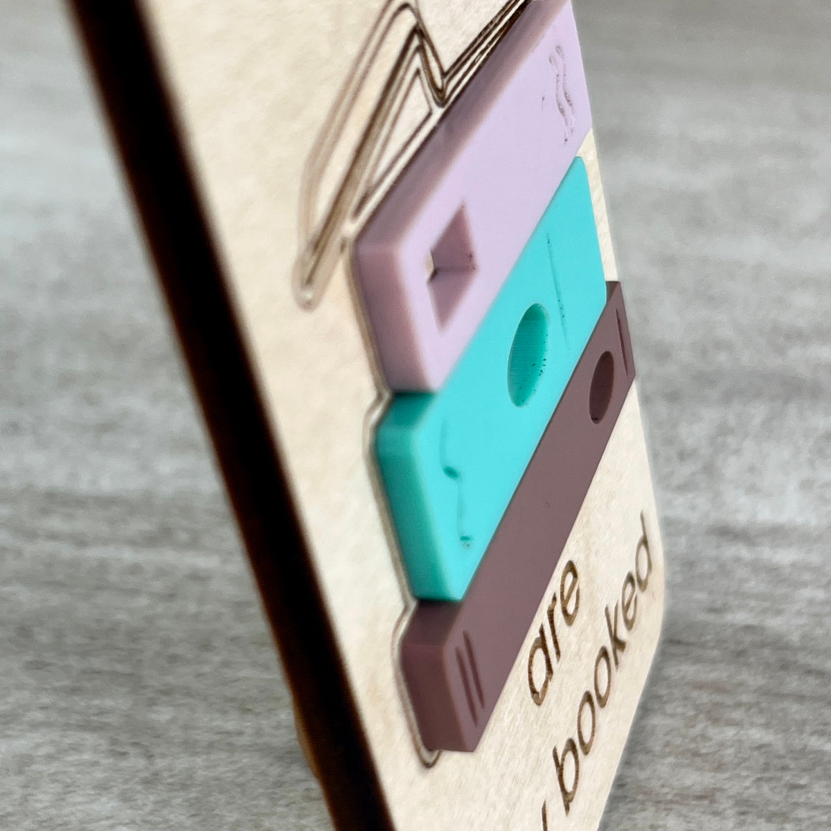 A close-up side view of a wooden magnet with a laser-cut acrylic layered design featuring three books in lavender, teal, and brown, highlighting the 3D effect of the acrylic element.