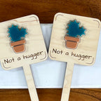 Two Not a Hugger maple wood plant stakes with teal acrylic cactus and terracotta pot side by side