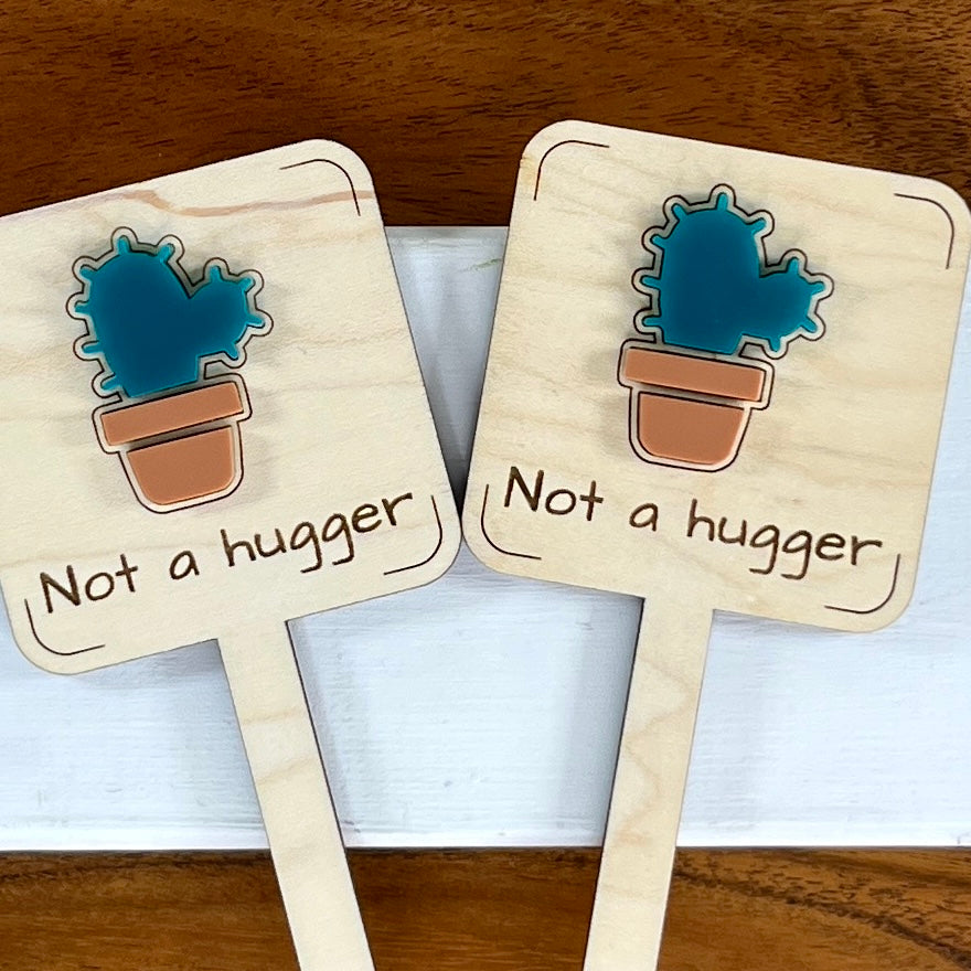 Two Not a Hugger maple wood plant stakes with teal acrylic cactus and terracotta pot side by side