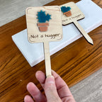 Hand holding two Not a Hugger laser-cut maple wood plant stakes with teal acrylic cactus