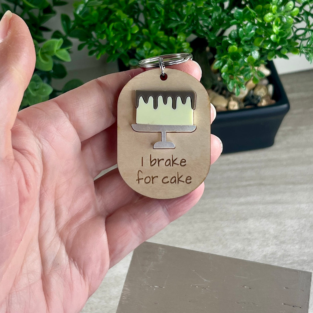 A hand holding an oval wooden keychain with a layered lemon acrylic drip cake and the engraving 'I brake for cake.'