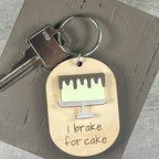 Oval wooden keychain with lemon acrylic drip cake, engraved 'I brake for cake,' attached to a metal key on a brown surface. 