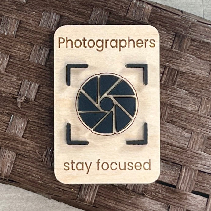 A rectangular wooden magnet laser-engraved with the words 'Photographers stay focused' and an acrylic camera aperture icon in the center, placed on a brown woven surface.