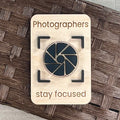 A rectangular wooden magnet laser-engraved with the words 'Photographers stay focused' and an acrylic camera aperture icon in the center, placed on a brown woven surface.