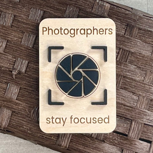 A rectangular wooden magnet laser-engraved with the words 'Photographers stay focused' and an acrylic camera aperture icon in the center, placed on a brown woven surface.