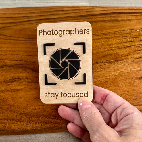 A hand holding a rectangular wooden magnet laser-engraved with the words 'Photographers stay focused' and a black acrylic camera aperture icon in the center, set against a wooden surface.