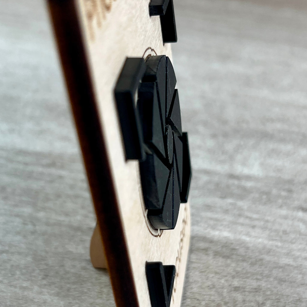 A side view close-up of a wooden magnet with a black camera aperture icon layer, standing on a light gray textured surface.