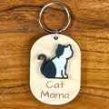 Oval wooden keychain with black and white acrylic cat, engraved 'Cat Mama,' on a wood surface with a metal split ring.