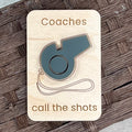 A rectangular wooden magnet with rounded corners featuring a grey acrylic whistle and the laser-engraved text 'Coaches call the shots'. The magnet is placed on a dark brown woven mat background.