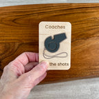 A hand holds a small wooden magnet engraved with the words 'Coaches call the shots' and featuring a dark gray acrylic whistle in the center, set against a polished wooden board background.