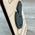 A side view close-up of a dark gray acrylic whistle on a light wood engraved magnet, set against a gray textured background and highlighting the 3D layering.