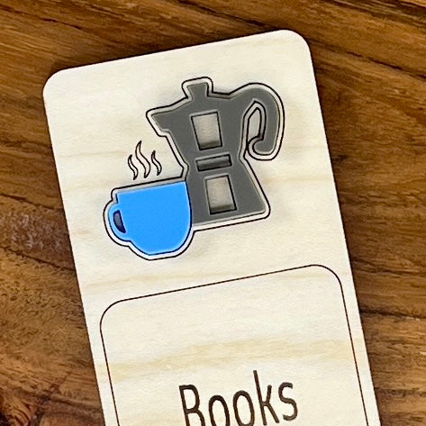 A close-up of a wooden book mark or bookmark labeled 'Books' with an acrylic mauve moka pot and blue coffee cup on a wood-grain background.
