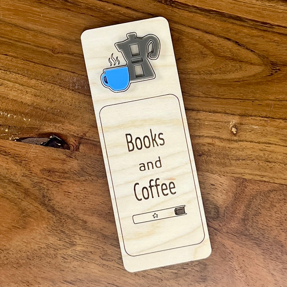 A rectangular wooden bookmark with rounded corners lies on a wooden tabletop. The bookmark is light natural wood with engraved text reading 'Books and Coffee' and small layered acrylic icons of a coffee pot and a blue coffee cup.