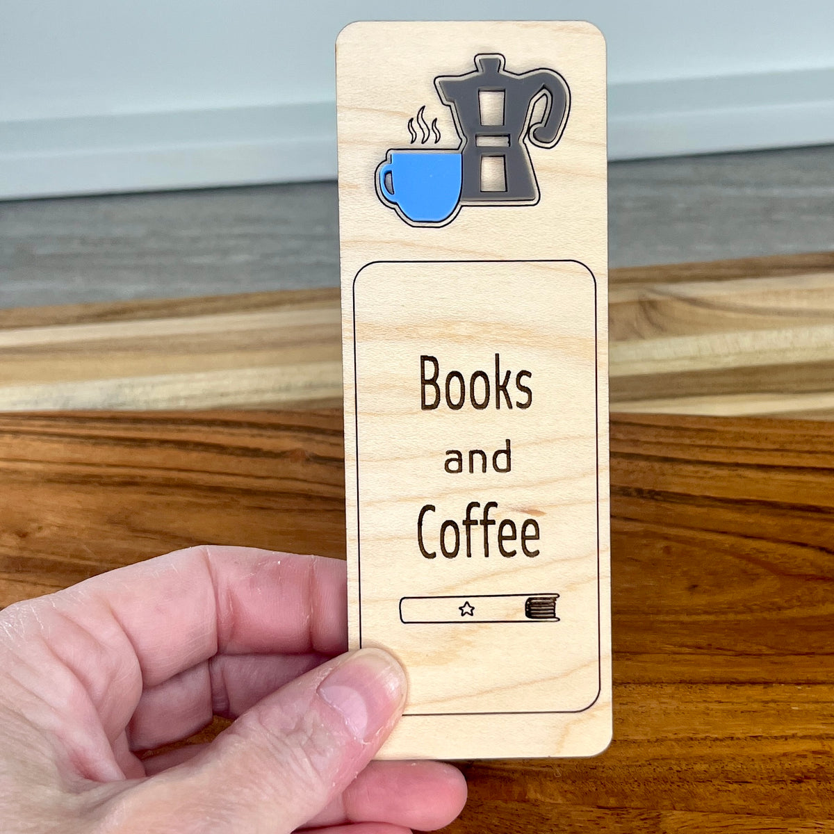 A hand holds a rectangular wooden book mark featuring a layered acrylic coffee mug and moka pot icon at the top and the words 'Books and Coffee' and a book engraved in a rounded rectangle in the center.