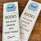Two wooden bookmarks with laser-engraved text and an acrylic grey armchair charm, displayed on a wooden surface. Both are laser-engraved with the message 'BOOKS because people are overrated'.