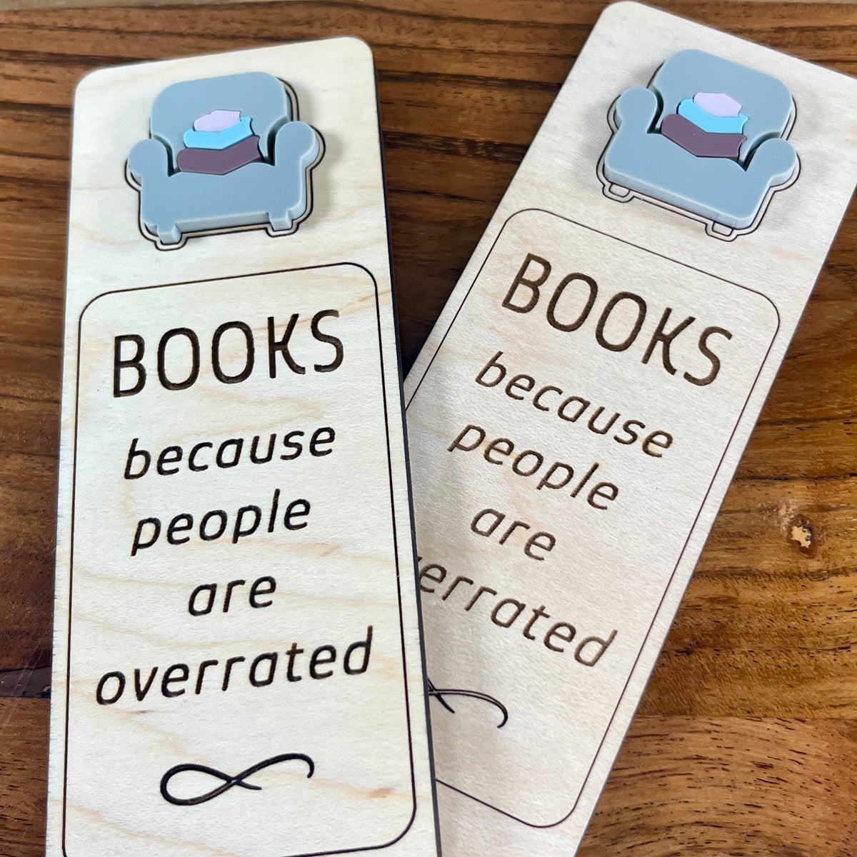 Two wooden bookmarks with laser-engraved text and an acrylic grey armchair charm, displayed on a wooden surface. Both are laser-engraved with the message 'BOOKS because people are overrated'.