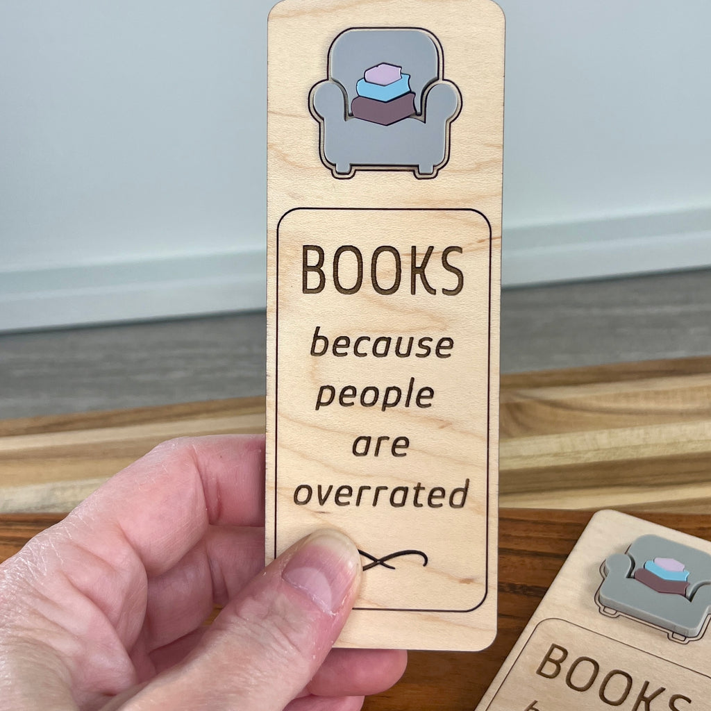 A hand holds a rectangular wooden bookmark laser-engraved with the phrase 'BOOKS because people are overrated' and a small acrylic armchair illustration. Another matching item is partially visible at the lower right on a wooden surface.