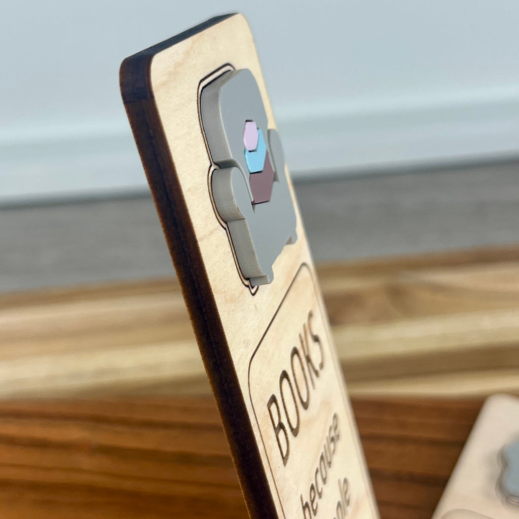 A side view of a wooden bookmark standing upright on a wooden surface, with a gray acrylic armchair and a stack of acrylic pastel coloured books, and the word 'BOOKS' engraved on the front.