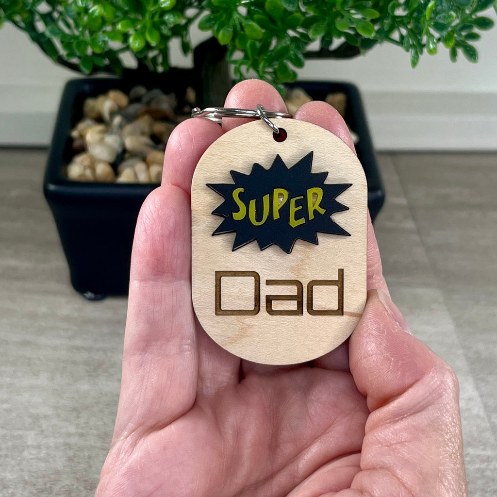 Hand holding Super Dad maple keychain with acrylic burst accent and key ring.