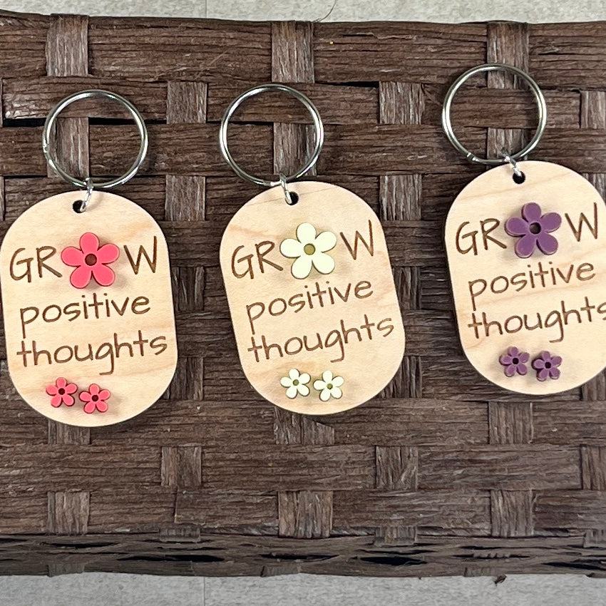 Multiple Grow Positive Thoughts maple keychains on display hooks showing purple, watermelon and lemon acrylic daisy colour variants
