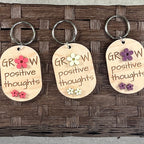 Multiple Grow Positive Thoughts maple keychains on display hooks showing purple, watermelon and lemon acrylic daisy colour variants