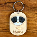 A wooden dog tag keychain engraved with 'Dog Mama' and featuring a black and white acrylic dog face design, shown on a wooden background.