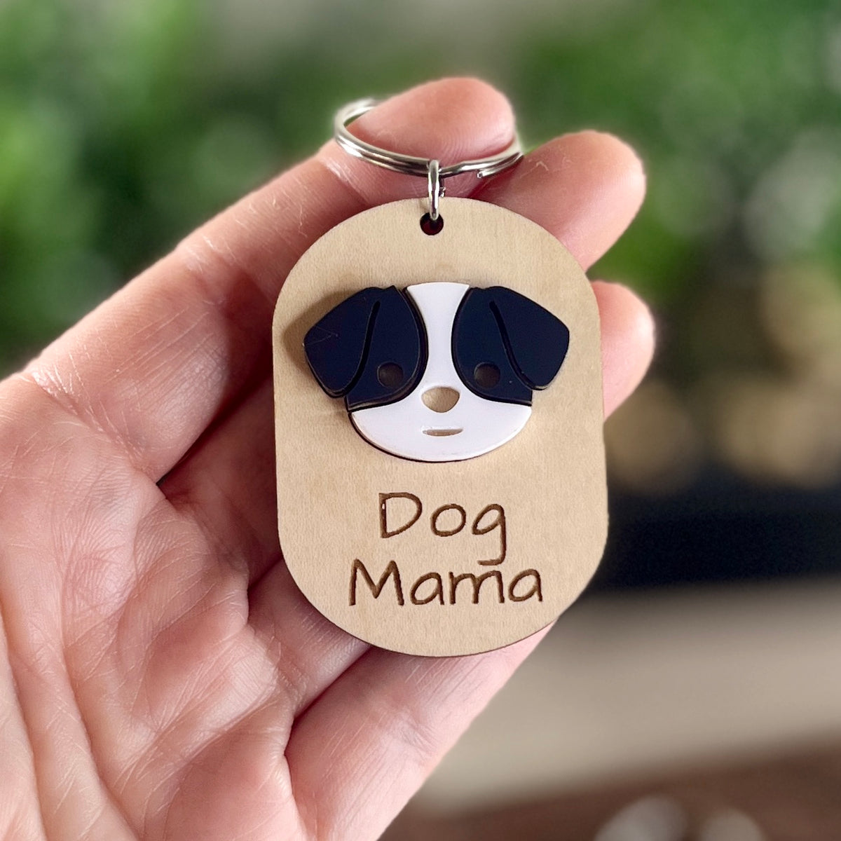 A hand holding a  wooden dog tag keychain featuring an acrylic dog face and the engraved text 'Dog Mama'.