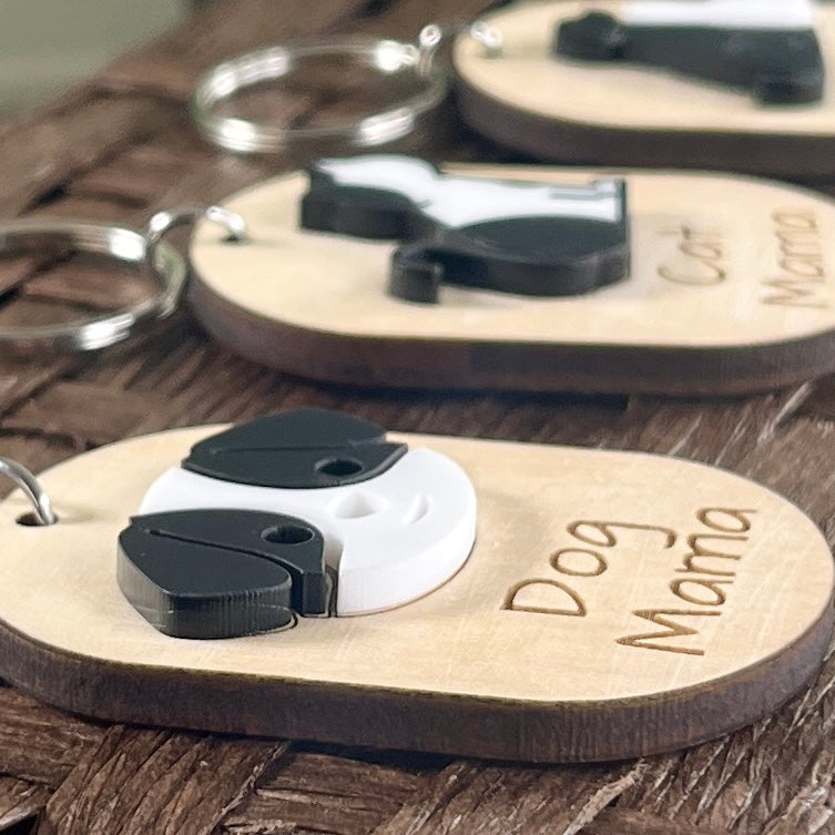 Three oval wooden keychains engraved with 'Dog Mama' arranged on a rustic wooden surface. The keychains feature a small 3D acrylic Dog designed to highlight the layered effect of the acrylic.