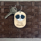 A wooden dog tag keychain with a laser-engraved 'Dog Mama' design and an acrylic dog face, attached to a metal keyring with a key. The keychain is displayed on a dark brown woven basket background.