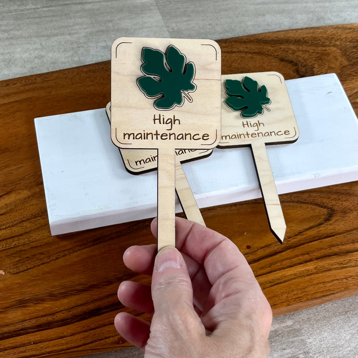 Hand holding two High Maintenance laser-cut maple wood plant stakes with forest green fig leaf