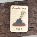 A rectangular wooden magnet with rounded corners featuring the engraved text 'Gardeners dig it' and a raised acrylic shovel in brown soil design. The magnet is photographed on a dark brown woven wicker surface.