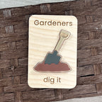 A rectangular wooden magnet with rounded corners featuring the engraved text 'Gardeners dig it' and a raised acrylic shovel in brown soil design. The magnet is photographed on a dark brown woven wicker surface.