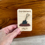 A hand holds a small wooden magnet engraved with the words 'Gardeners dig it' around an acrylic shovel illustration, against a wooden tabletop background.