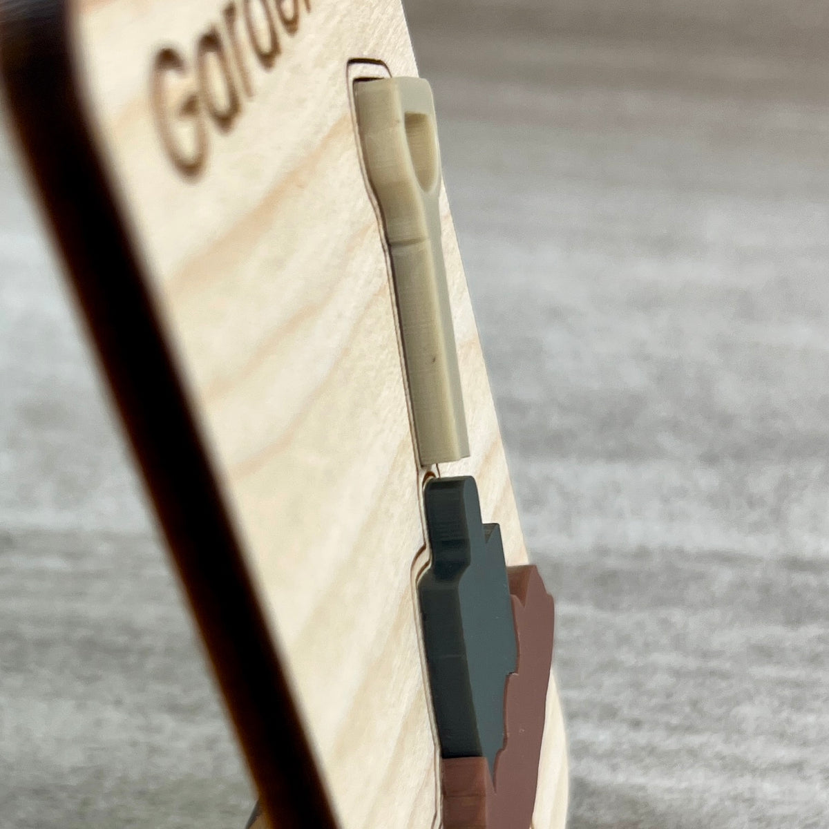 Side view close-up of a wooden magnet with a layered acrylic shovel in soil design, photographed on a gray textured surface and designed to highlight the 3D effect from the acrylic.