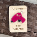 A pink and black acrylic glue gun with daisy cutouts, mounted on a light wood magnet that reads 'Crafters see potential,' placed on a brown woven surface.