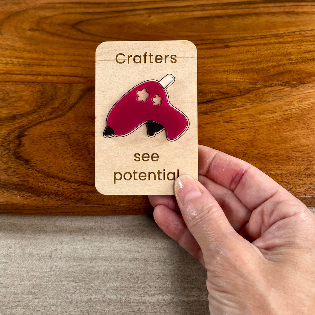 A hand holds a small wooden magnet featuring a pink and black acrylic glue gun with the words 'Crafters see potential' laser-engraved on it, set against a wooden tabletop background.