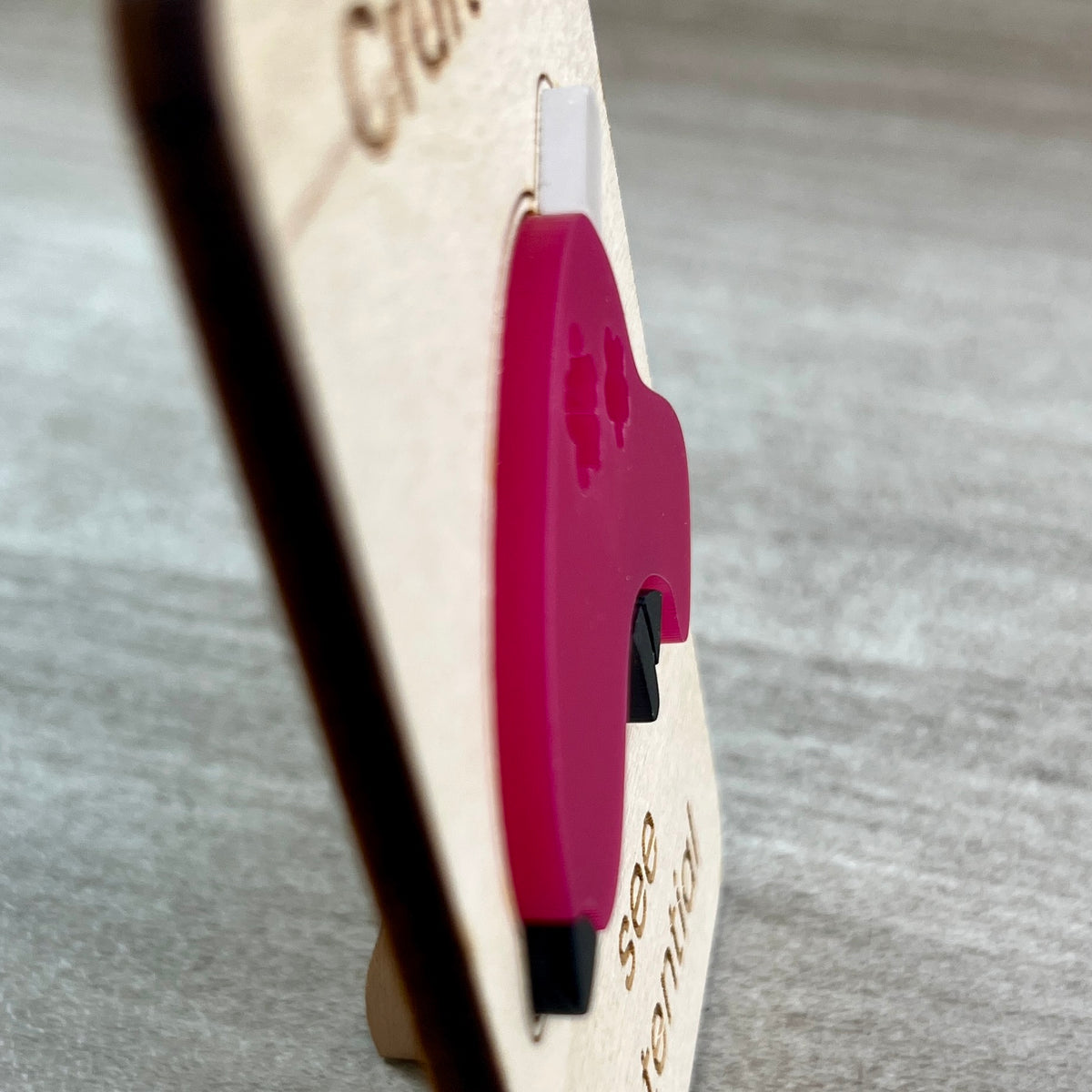A side view close-up of a pink acrylic glue gun layered on a wooden magnet, photographed on a gray textured surface and designed to highlight the 3D feature of the product.