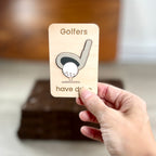 A hand holds a small wooden magnet that reads "Golfers have drive" with a simple golf club and ball illustration. The background is softly blurred, showing an indoor setting with a wooden surface.
